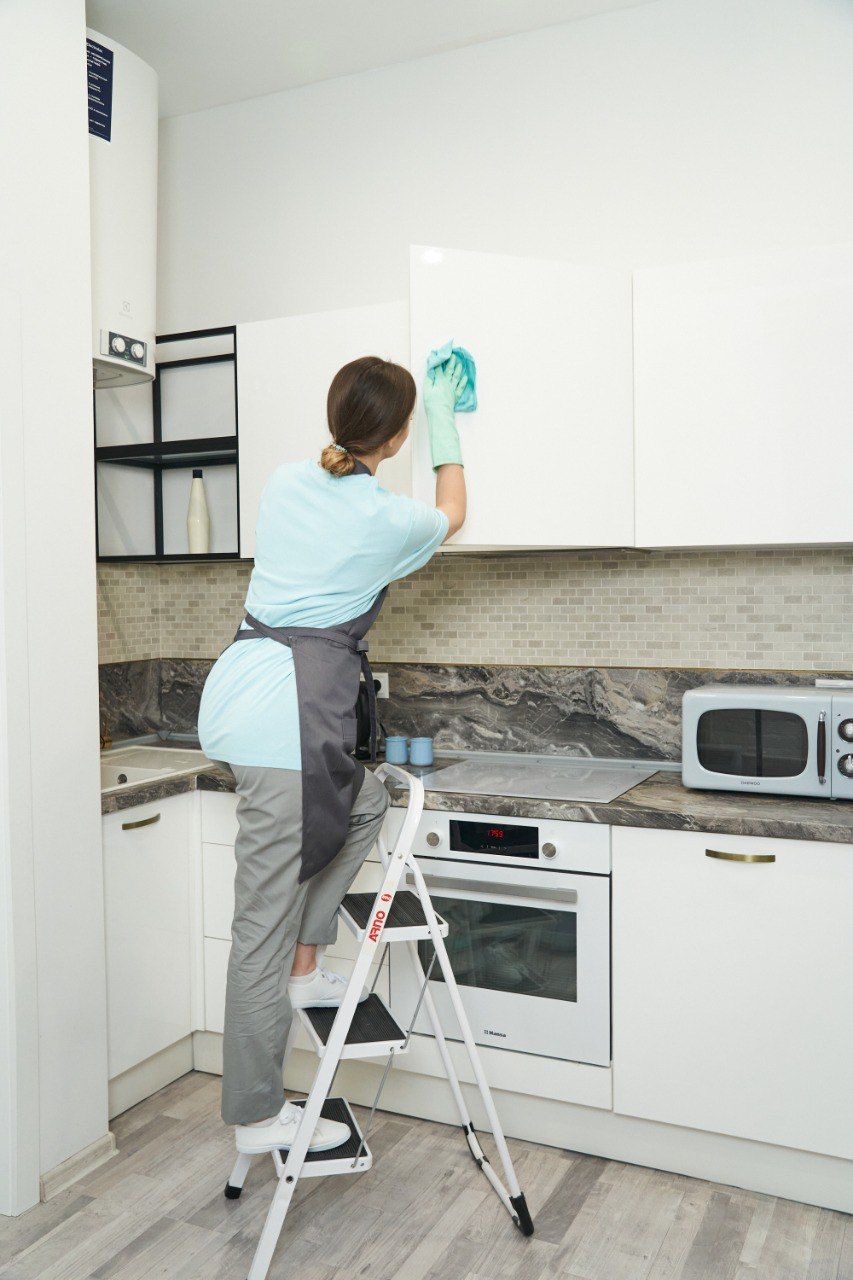 Cleaner on a step stool wiping white kitchen cabinets with a blue cloth